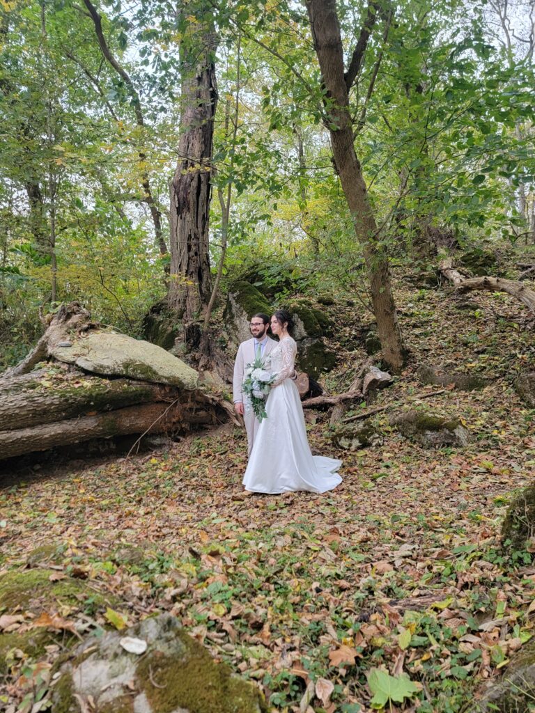 Matthew and Abigail standing at the rustic entrance of The Cove at Timber Valley Farm Barn Rental Wedding and Event Venue, framed by lush greenery and fallen leaves.