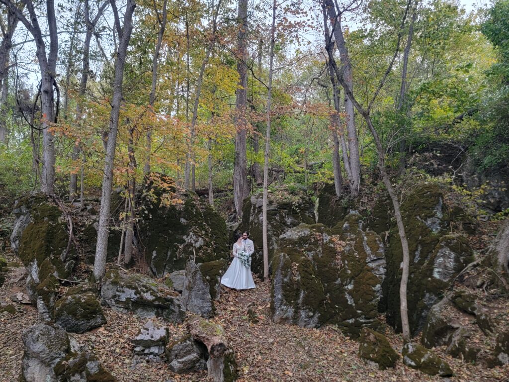 A wide-angle distance shot of Abigail in her flowing white wedding dress, surrounded by massive moss-covered boulders within the natural woodland of The Cove at Timber Valley Farm Barn Rental Wedding and Event Venue.