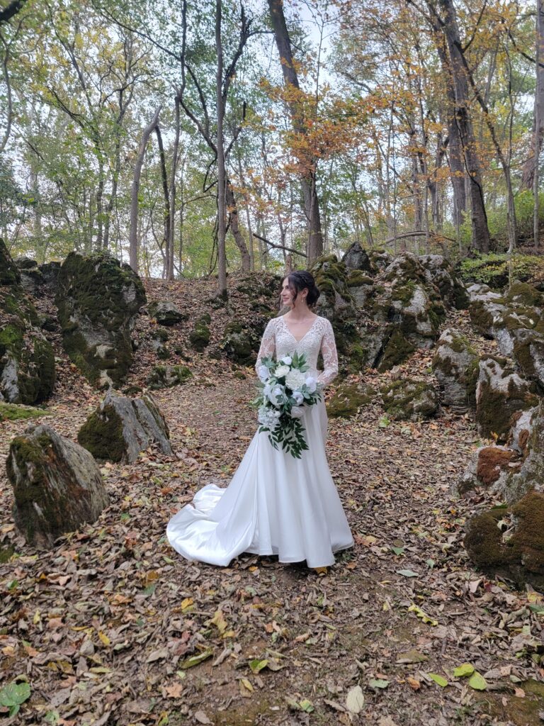 A view of Abigail in her white wedding gown standing among massive moss-covered boulders in the wooded area of The Cove at Timber Valley Farm Barn Rental Wedding and Event Venue.