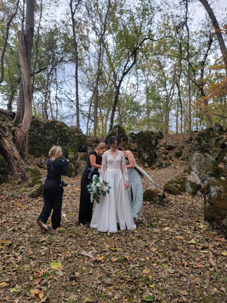 Photo Gallery- A behind-the-scenes shot of photographers and Abigail’s sister assisting the bride with her gown among the fallen October leaves at The Cove within Timber Valley Farm Barn Rental Wedding and Event Venue.