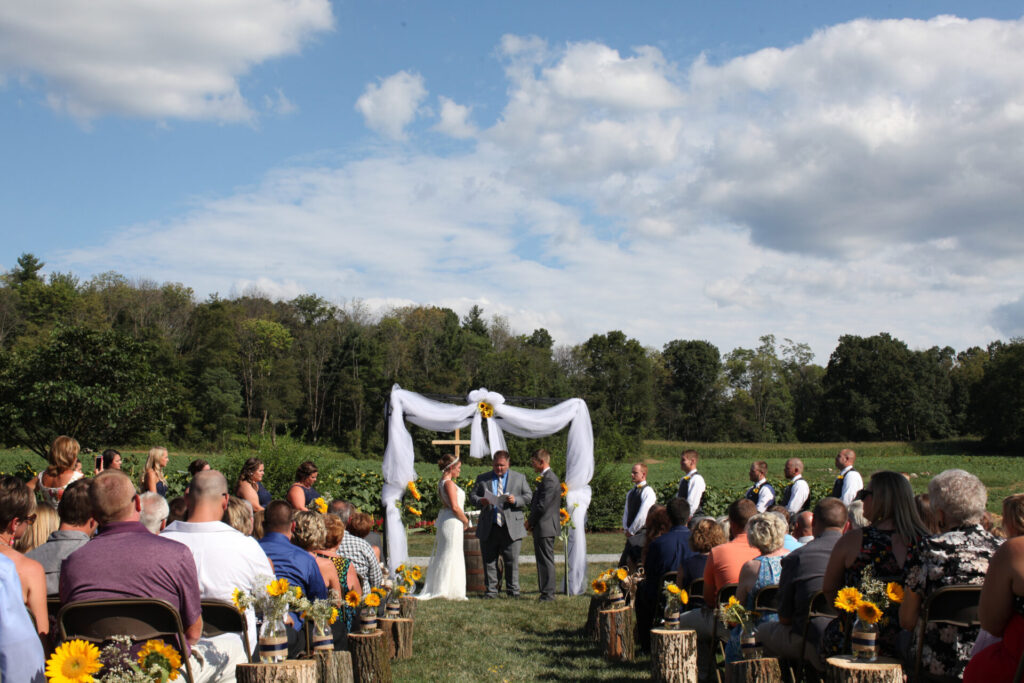 Bride and groom exchanging vows beneath the Wedding Arch with The Cross in the valley and sunflower field at Timber Valley Farm Barn Rental in Clear Spring, Maryland.