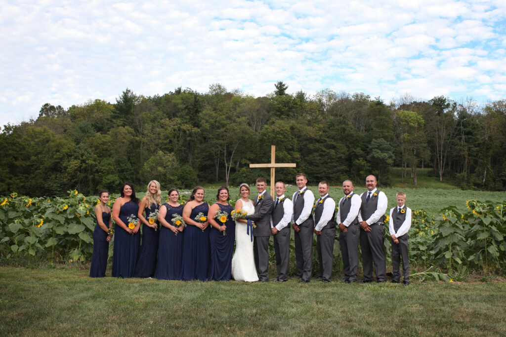 Bride, groom, and wedding party posing beside The Cross surrounded by blooming sunflowers in the valley at Timber Valley Farm Barn Rental in Clear Spring, Maryland.