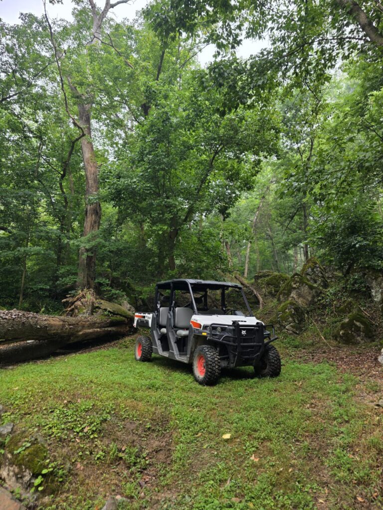 A Bobcat utility vehicle parked near God's Cathedral at Timber Valley Farm Barn Rental, ready to transport the once-forgotten gate, tools, and supplies to its new home in the peaceful woodland setting of God's Cathedral Maryland.