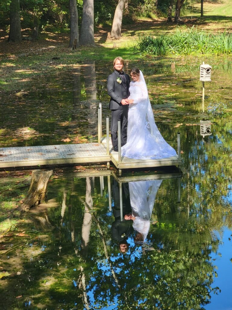 Bride and groom standing on the dock overlooking the Spring-Fed Pond with beautiful reflections at Timber Valley Farm Barn Rental in Clear Spring, Maryland.