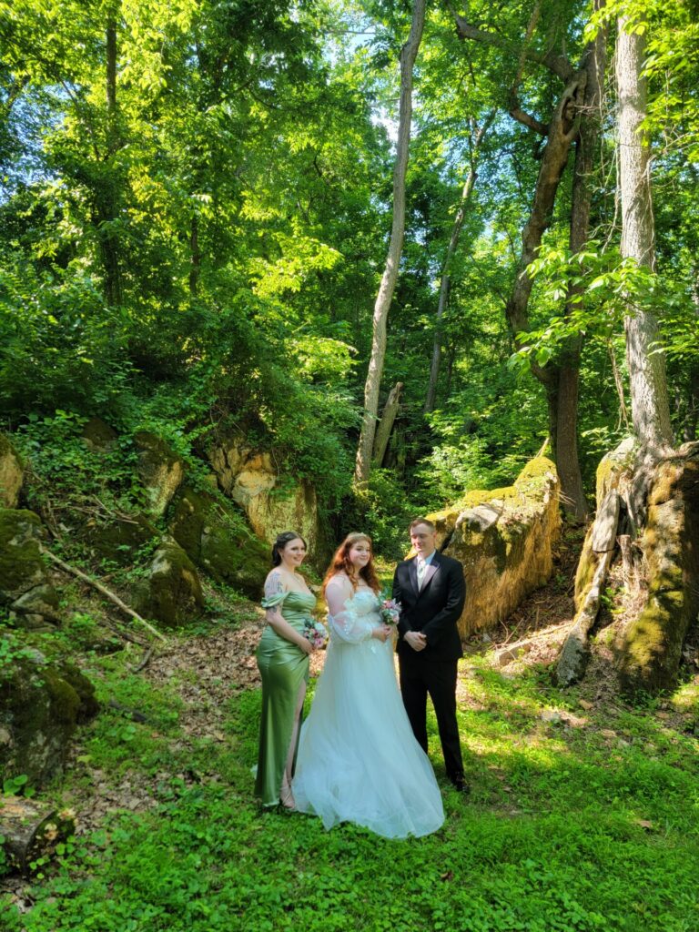 Bride, groom, and bridesmaid surrounded by towering trees and lush woodland scenery in the Enchanted Forest at Timber Valley Farm Barn Rental in Clear Spring, Maryland.