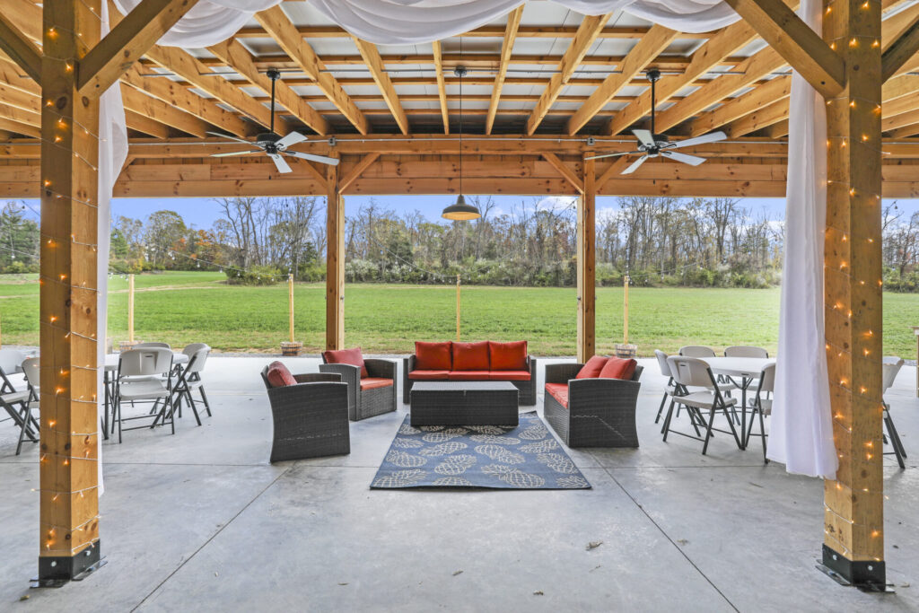 Covered outdoor wedding space inside the open-sided timber-frame barn with seating, string lights, ceiling fans, and views across the lawn at Timber Valley Farm Barn Rental in Clear Spring Maryland.