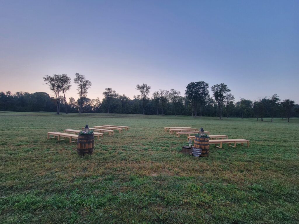 Open countryside wedding ceremony setting with wooden benches arranged across a grassy field surrounded by mature trees at Timber Valley Farm Barn Rental in Clear Spring Maryland.