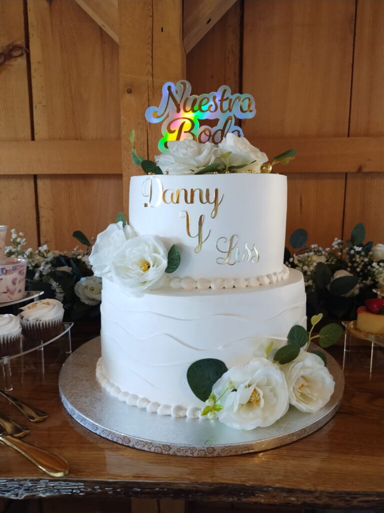A personalized wedding cake featuring the couple's names, a “Nuestra Boda” topper, and white floral accents at Timber Valley Farm Barn Rental.