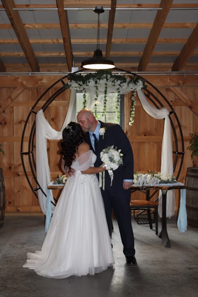 Intimate indoor wedding ceremony inside the timber-frame barn at Timber Valley Farm Barn Rental in Clear Spring Maryland, with the couple standing beneath a ceremony arch.