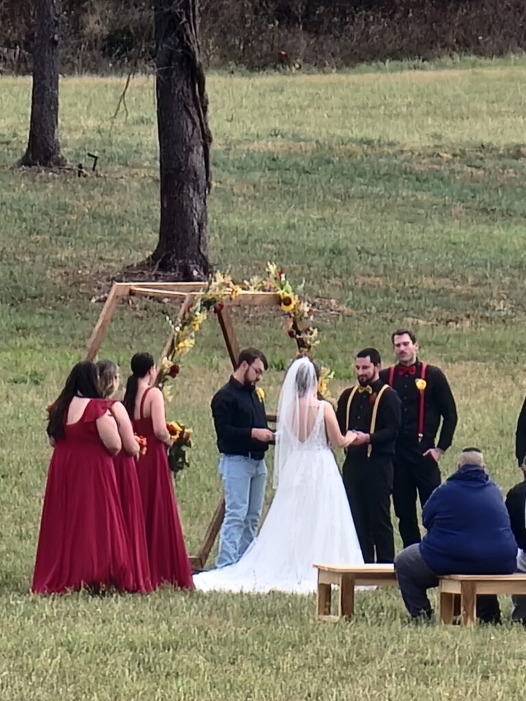 Simple outdoor wedding ceremony backdrop with wooden arbor, seasonal flowers, bride, groom, and wedding party at Timber Valley Farm Barn Rental in Clear Spring Maryland.