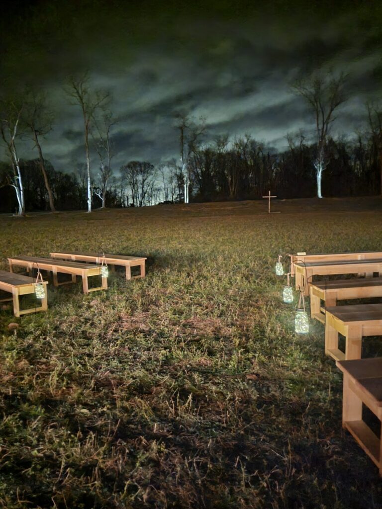 Evening wedding ceremony backdrop with illuminated lanterns, wooden benches, open field, trees, and cross at Timber Valley Farm Barn Rental in Clear Spring Maryland.