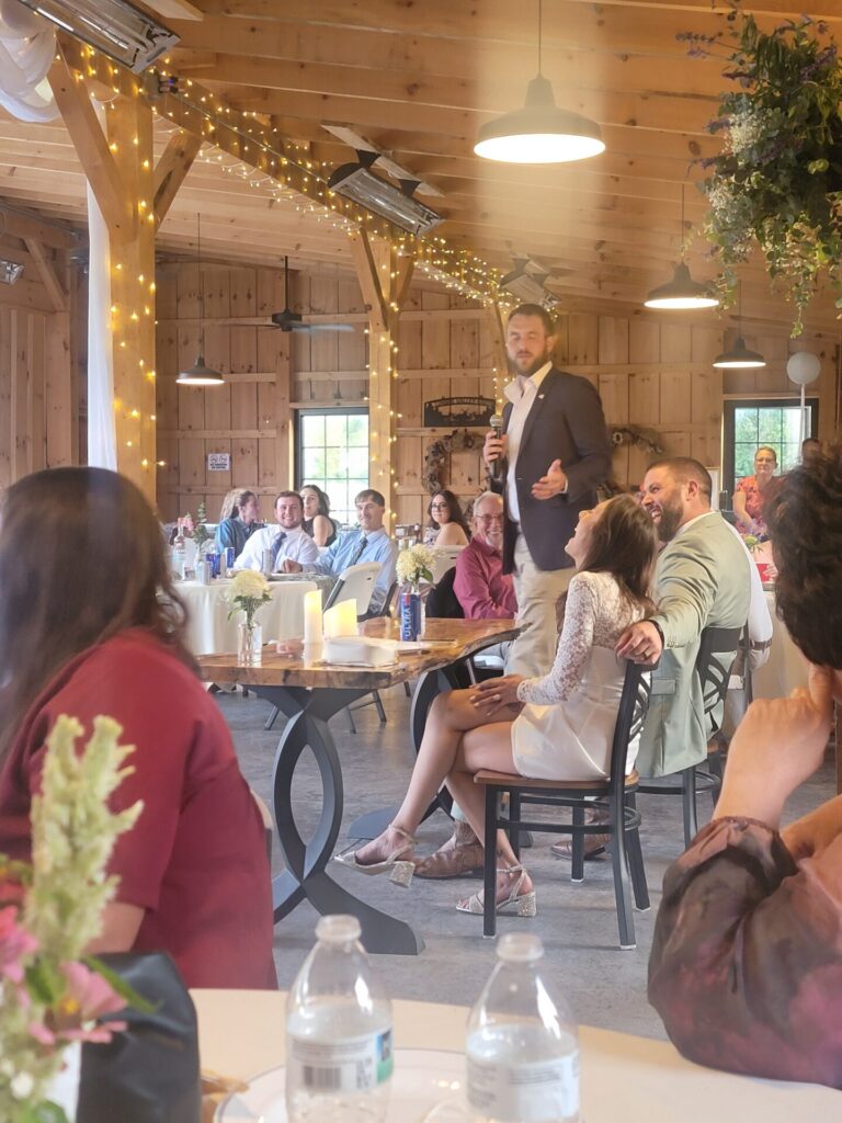 Wedding guests gather around tables inside the timber-frame barn at Timber Valley Farm Barn Rental, with the open patio and surrounding property nearby.