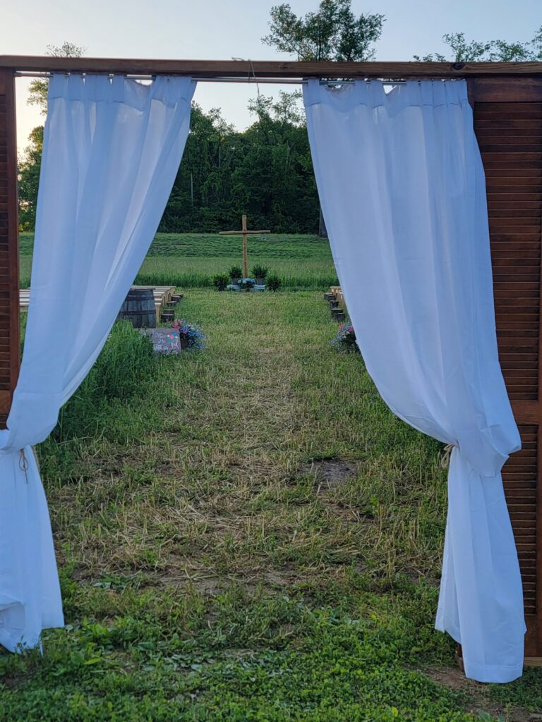 Outdoor wedding backdrop ideas featuring a wooden backdrop with flowing white drapery framing the ceremony aisle and cross at Timber Valley Farm Barn Rental in Clear Spring, Maryland.