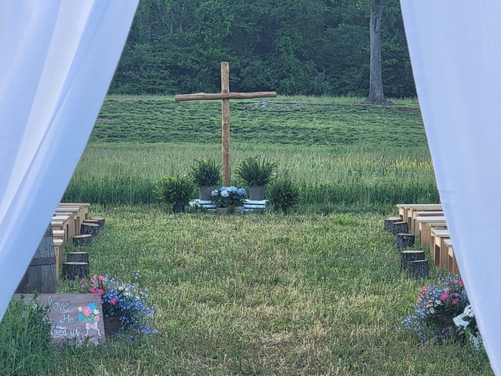 Outdoor Wedding Ceremony setting viewed through flowing white fabric, showing a wooden cross, ceremony benches, open lawn, and surrounding trees at Timber Valley Farm Barn Rental in Clear Spring Maryland.