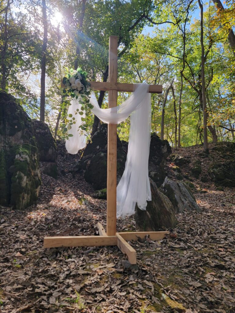 Rustic Wedding Backdrop with a decorated wooden cross, flowing fabric, greenery, moss-covered rocks, and mature trees at Timber Valley Farm Barn Rental in Clear Spring, Maryland.
