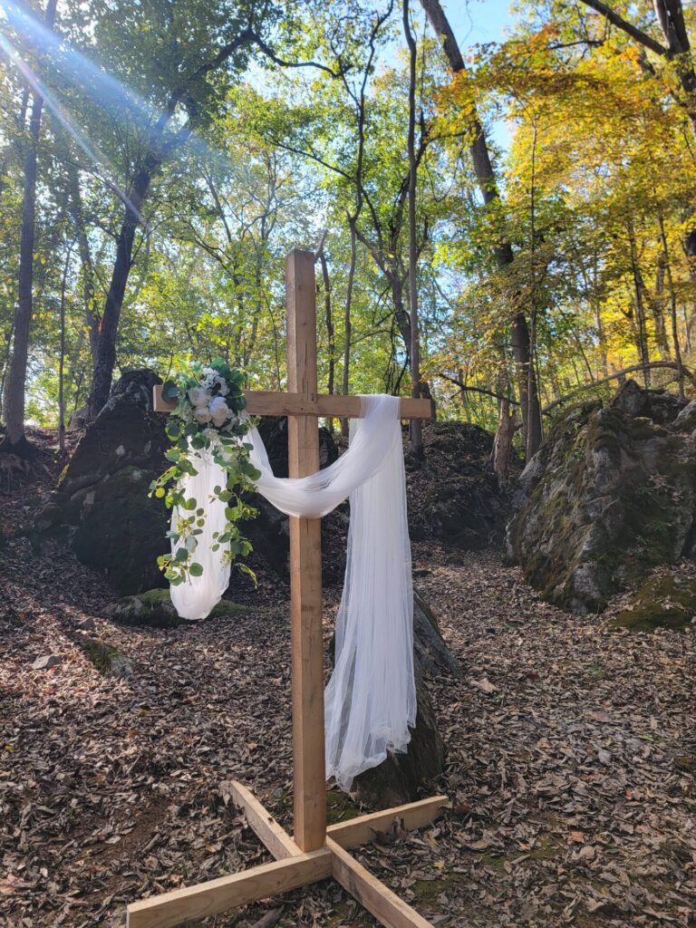 Wedding Ceremony Decor featuring a decorated wooden cross with flowing fabric and greenery in a natural woodland setting at Timber Valley Farm Barn Rental in Clear Spring Maryland.