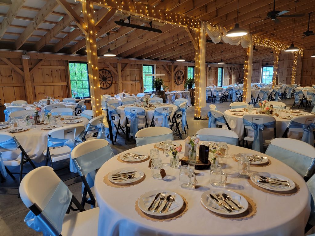 Maryland Barn Wedding. Rustic timber-frame barn wedding reception with round guest tables, blue chair sashes, and warm string lights at Timber Valley Farm Barn Rental in Clear Spring, Maryland